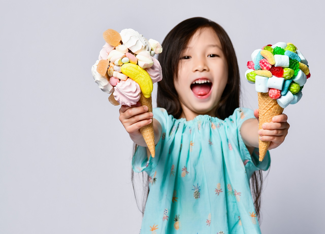 uśmiechnięta dziewczynka trzymająca dwie porcje kolorowych lodów dla dzieci z dodatkami: żelkami i piankami